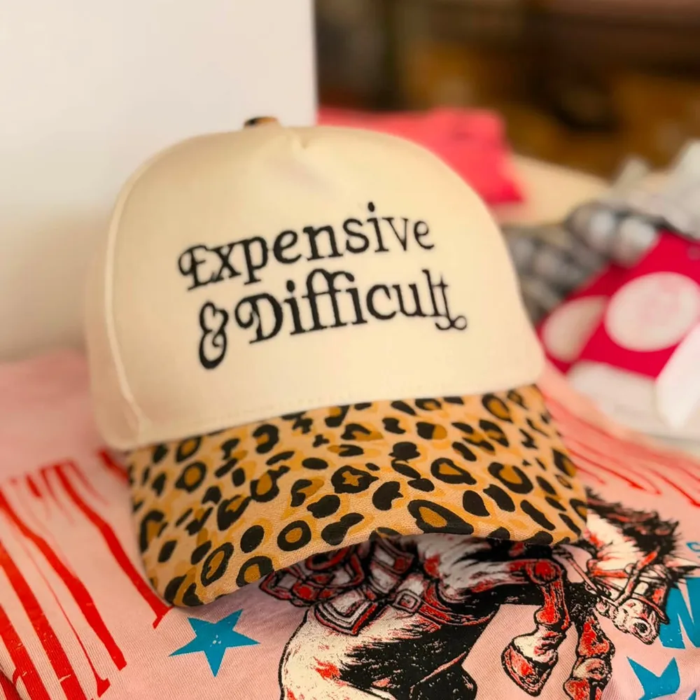Cream trucker cap with a leopard print brim, embroidered in ornate black type: Expensive and Difficult.