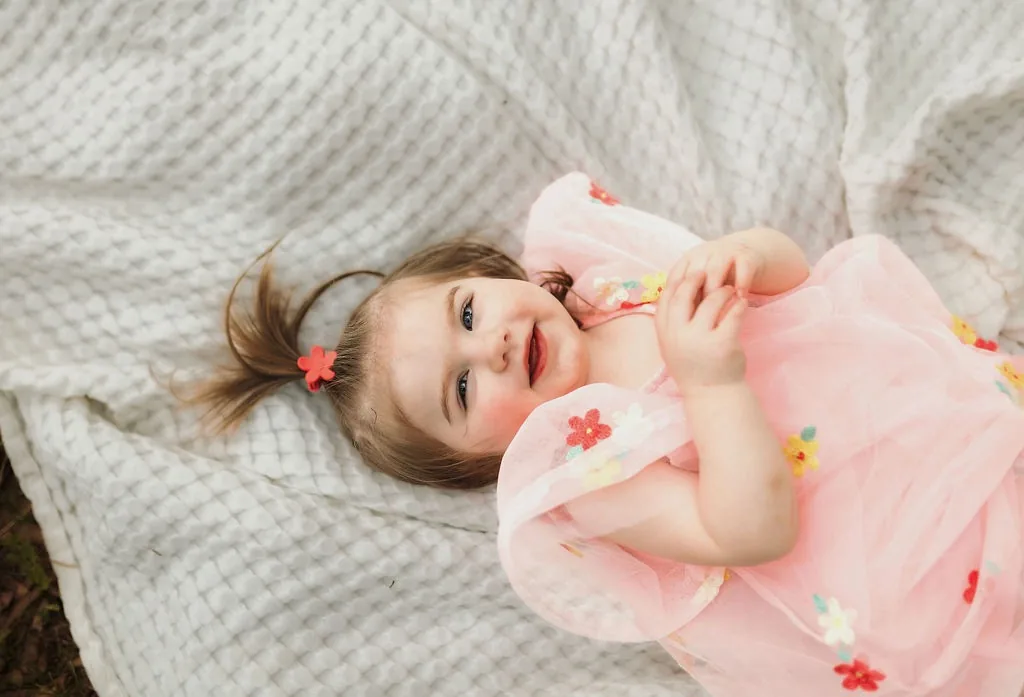 Lindie Sue as a toddler in a pink floral tulle dress, lying on a white waffle blanket and smiling up at the camera.