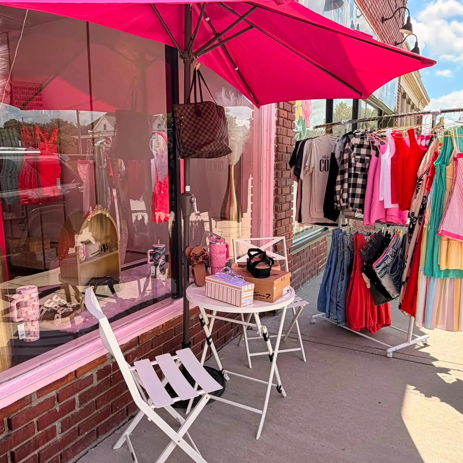 Lindie Sue's storefront: a hot pink umbrella over a white bistro table, a rack of clothes on the sidewalk, pink window trim on brick.